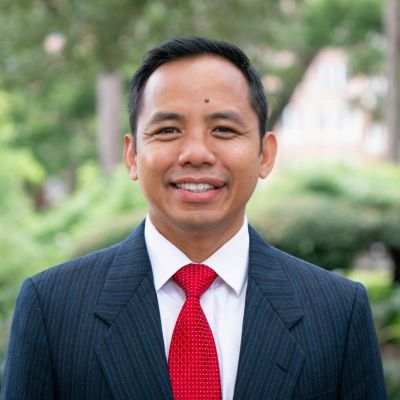 A man in a suit and tie, standing outdoors, he is smiling